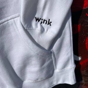 May include: White hoodie with a kangaroo pocket and a drawstring hood. The left sleeve features the embroidered word "w;nk" in black. The hoodie is made of a soft, comfortable material.