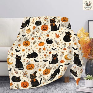 May include: A cream-colored throw blanket featuring a repeating pattern of black cats, orange jack-o'-lanterns, bats, crescent moons, and autumn leaves. Some cats wear orange scarves or witch hats, and string lights connect several of the pumpkins.