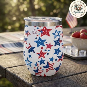 May include: A white insulated tumbler with a clear lid, decorated with a pattern of red and blue stars. Some stars feature stripes, resembling the American flag. The tumbler is designed for cold beverages.