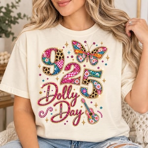 May include: A cream-colored t-shirt featuring a graphic design with the text 925 Dolly Day. The design includes a colorful butterfly, a small guitar, and numbers filled with patterns of leopard print, pink gingham, and floral motifs in bright colors.