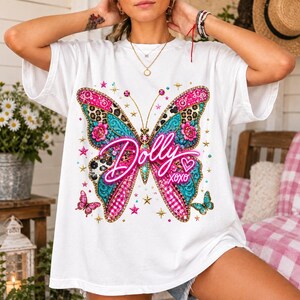 May include: A white short-sleeve t-shirt featuring a large, colorful butterfly graphic on the front. The butterfly wings display patterns of leopard print, pink floral motifs, and turquoise textures. The text Dolly XOXO is written in pink.