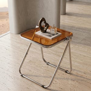 Puede incluir: Una mesa auxiliar moderna con una tapa transparente de color ámbar y un marco cromado pulido. La mesa contiene una escultura de bronce, un libro y un frasco de perfume. La mesa cuadrada tiene un diseño minimalista.