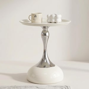 Peut inclure: Une table d'appoint blanche avec un plateau rond et une base chromée en forme de sablier. Une tasse beige avec une illustration de femme et un bougeoir blanc en forme de nuage sont posés sur le plateau. La table est une pièce d'accent moderne.
