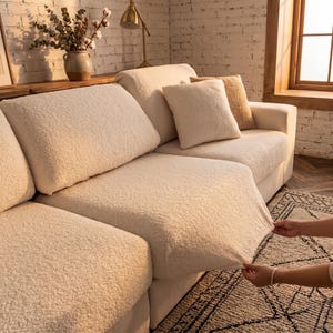 May include: Cream-coloured, textured sofa cover being stretched over a sectional sofa. The sofa has several matching throw cushions in cream and tan. The background includes a brick wall, a wooden shelf with a vase, and a window.