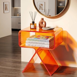 Op de afbeelding: Een moderne, transparante oranje bijzettafel met een uniek geometrisch ontwerp. De tafel heeft een plank met boeken zoals "Sculpture & Space" en decoratieve items. De tafel is gemaakt van een helder, oranje materiaal en heeft een eigentijdse uitstraling. Afmetingen in centimeters.