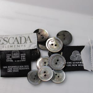 May include: A collection of round, grey, two-hole buttons featuring the Escada brand name embossed on the surface. Included are two black fabric garment labels with white text, one reading Escada Elements and the other Pure New Wool.