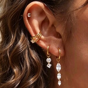 May include: A gold ear cuff with a clear stone, a gold stud with a clear stone, and two gold hoop earrings with clear stone dangles. One hoop features a round and square stone, while the other features three marquise-shaped stones in a vertical line.