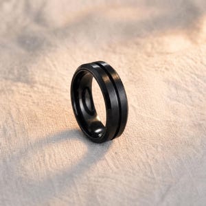 Op de afbeelding: Een matzwarte ring met een gladde, geborstelde afwerking en een enkele, dunne, gepolijste groef die horizontaal rond het midden van de band loopt. De ring heeft een afgeschuind randontwerp, wat zorgt voor een opvallend profiel met een donkere kleur.