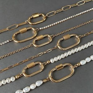May include: A collection of gold-toned necklaces featuring oval carabiner-style clasps. The chains include a mix of elongated links, a delicate rhinestone strand, a fine cable chain, and a strand of white freshwater pearls.
