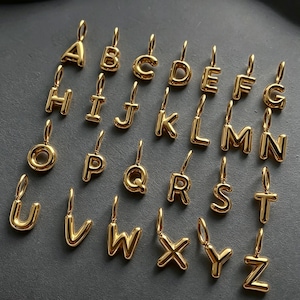 Puede incluir: Una colección de veintiséis dijes de letras de metal en tono dorado, cada uno con un diseño liso, redondeado y abombado, con una pequeña argolla en la parte superior para colgar. Las letras del alfabeto están dispuestas de la A a la Z.