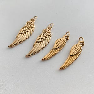 May include: Four gold-toned metal charms shaped like angel wings, each featuring a small circular loop at the top for attachment. The charms display detailed, embossed feather textures along the surface, with a polished finish and a curved design.