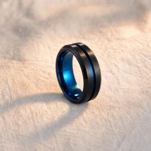 May include: A ring featuring a matte black exterior with a brushed finish and a thin, centred groove that reveals a polished, vibrant blue interior. The band has bevelled edges and a smooth, rounded profile, showcasing a contrast between the dark outer surface and the bright blue inner lining.