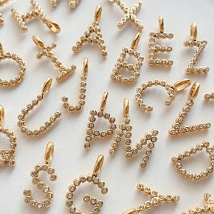 Pode incluir: Uma coleção de berloques de letras em metal dourado, cada um apresentando uma série de pequenos cristais circulares transparentes incrustados. Estes pingentes de alfabeto são projetados com uma pequena alça no topo para fixação, exibindo várias letras, incluindo A, B, E, G, J, M, P, S e Z, em uma construção uniforme.