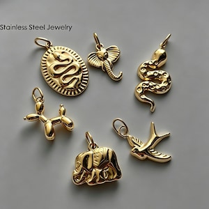 Puede incluir: Una colección de seis dijes de acero inoxidable en tono dorado que incluyen un colgante de serpiente ovalado, una cabeza de elefante, una serpiente enroscada, un perro globo, un elefante caminando y un pájaro en vuelo. Texto Stainless Steel Jewelry.