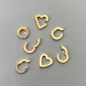 Può includere: Una collezione di sette chiusure per gioielli in metallo color oro di varie forme. Due sono a forma di cuore, tre sono circolari e due sono circolari con una fila di piccole pietre trasparenti incastonate su un lato. Ogni pezzo presenta un meccanismo di apertura a cerniera.