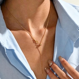 Peut inclure: Un collier lariat doré composé d'une chaîne fine, d'un petit fermoir circulaire et d'une perle coulissante permettant d'ajuster la longueur. Le collier se termine par un pendentif en forme de fine barre métallique droite à la base.