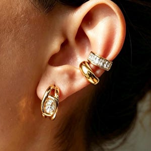 May include: A gold-toned earring set consisting of a hoop earring with a central oval clear stone and two ear cuffs. One ear cuff is a smooth polished gold band, while the other features a row of rectangular clear stones set in a gold band.