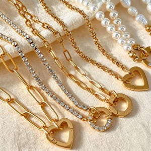 Puede incluir: Una colección de collares dorados con varios estilos de cadena, incluyendo una cadena tipo tenis con cristales, una cadena de eslabones ovalados gruesos y una hilera de perlas blancas. Varios collares tienen cierres dorados en forma de corazón.