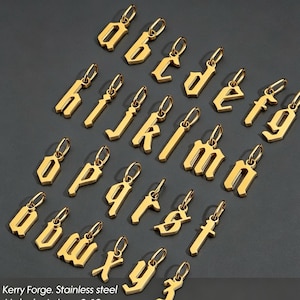 May include: A collection of gold-colored stainless steel letter pendants featuring lowercase characters in an old English font style. Each individual letter charm includes a small circular jump ring at the top for attachment to a necklace chain.