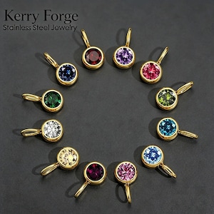 May include: A collection of twelve Kerry Forge stainless steel jewelry pendants arranged in a circle. Each pendant features a round, faceted gemstone in a different color, including red, purple, pink, green, blue, and clear, set in gold-tone metal.