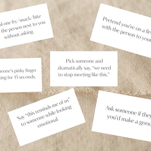 May include: White rectangular cards with black text, each featuring a different conversation starter. The cards are scattered on a beige fabric surface. The text prompts playful interactions, such as "Steal one fry..." and "Pretend you're on a first date..."