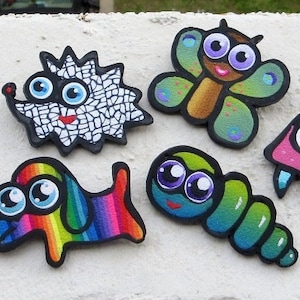 Könnte beinhalten: Fünf handbemalte Holz-Pins mit Comicfiguren. Die Figuren umfassen einen Igel, einen Schmetterling, eine Raupe, einen Hund und eine Qualle. Die Pins sind alle bunt und haben große, ausdrucksstarke Augen.