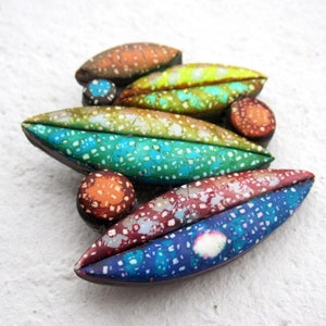 May include: A colorful abstract brooch made of polymer clay. The brooch features a cluster of leaf-shaped pieces in shades of green, blue, red, orange, and yellow. The pieces are decorated with white speckles and have a glossy finish.