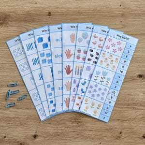 Könnte beinhalten: Ein Set aus sieben pädagogischen Zählstreifen mit blauem Rand, die Illustrationen von Würfeln, Strichlisten, Blöcken, Händen, Luftballons, Cupcakes, Kerzen, Geschenken, Partyhüten, Vögeln, Schnecken und Blumen sowie den Text Wie viele zeigen.