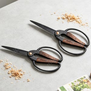 May include: Two pairs of black metal scissors with dark wood handle inlays. One pair is engraved with the name Sophia and the other with the name Lucas. The scissors feature large oval finger loops and a central copper-colored pivot screw.