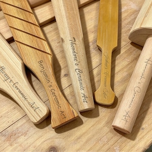 May include: A collection of light-coloured wooden pottery tools with handles. Each tool is engraved with a name followed by the text Ceramic Art. One tool features diagonal grooves on its surface, while others have smooth, cylindrical or flat shapes.
