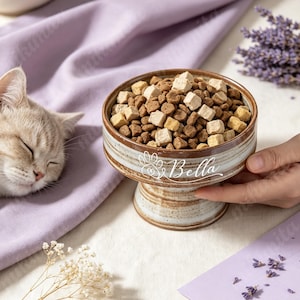 以下が含まれることがあります： 茶色とクリーム色の釉薬仕上げが施された陶器製の脚付きペット用ボウル。中にはドライフードが入っています。ボウルには白い筆記体で「Bella」という名前が刻まれており、その横には中央に小さなハートがあしらわれた白い肉球のアイコンが描かれています。