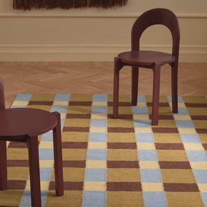 May include: Two dark maroon chairs with curved backrests and four straight legs, positioned on a rectangular area rug featuring a repeating grid pattern of light blue, mustard yellow, and dark brown squares across the entire surface.