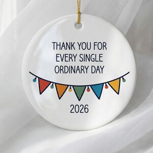May include: A round white ceramic ornament hanging from a gold string. The text reads THANK YOU FOR EVERY SINGLE ORDINARY DAY above a colorful bunting banner with triangular flags in red, orange, green, and blue, followed by the year 2026 below.