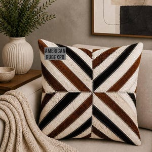 May include: Square decorative pillow with a geometric pattern of brown, black, and cream-colored stripes. The pillow is on a beige sofa, with a beige throw blanket and a vase with greenery in the background. The text "AMERICAN RUGEXPO" is visible on the pillow.