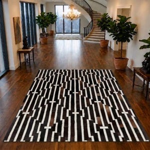 Op de afbeelding: Een rechthoekig vloerkleed met een herhalend geometrisch patroon van afwisselende zwarte, witte en donkerbruine verticale strepen. Het kleed is samengesteld uit gestikte panelen, wat zorgt voor een getextureerd oppervlak met een contrastrijk lineair ontwerp.