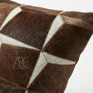 May include: A square throw pillow cover crafted from panels of genuine cowhide fur in shades of dark brown, medium brown, and cream. The pieces are stitched together in a geometric pattern. A logo with the letters A and H is visible on the front.