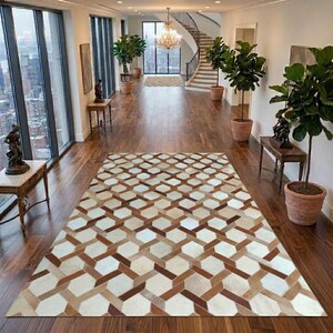 Op de afbeelding: Een rechthoekig vloerkleed met een geometrisch patroon van in elkaar grijpende zeshoeken in tinten crème, beige en donkerbruin. Het kleed ligt op een donkere houten vloer in een ruime hal met grote ramen, kamerplanten en een trap.