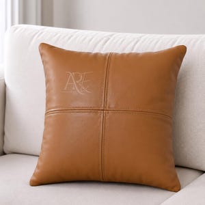 May include: A square tan leather throw cushion featuring a cross-stitched seam design that divides the front surface into four equal quadrants. A stylised logo with the letters AE is embossed in the upper left quadrant of the cushion.