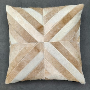May include: A square throw pillow featuring a geometric pattern of alternating diagonal stripes in shades of tan, beige, and cream. The surface is constructed from stitched panels of hair-on hide, creating a textured, multi-tonal appearance.