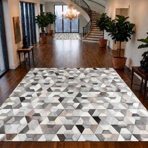 Op de afbeelding: Een groot rechthoekig vloerkleed met een geometrisch patroon bestaande uit diverse tinten grijs, wit en zwart in zeshoekige en driehoekige vormen. Het kleed heeft een getextureerd oppervlak met een herhalend mozaïekontwerp over het hele vloeroppervlak.