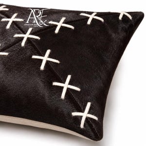 May include: A black cowhide pillow featuring a geometric pattern of white cross-stitch detailing. The surface has a textured hair-on-hide finish. The logo for American Rug Expo is printed in white in the upper left corner of the pillow cover.
