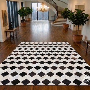 Op de afbeelding: Een rechthoekig vloerkleed met een herhalend geometrisch patroon van zwarte en witte ruiten. Het kleed ligt plat op een donkere houten vloer in een grote kamer met hoge plafonds, grote ramen en verschillende groene planten.