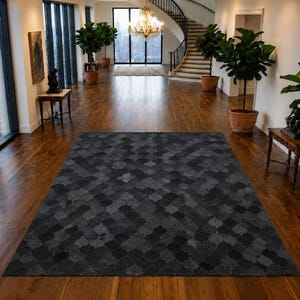 Puede incluir: Una alfombra rectangular con un patrón geométrico en gris carbón oscuro y negro compuesto por formas de cuatrifolio entrelazadas. La alfombra tiene una textura de pelo corto y está colocada en el centro sobre un suelo de madera pulida.