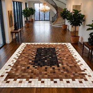 Könnte beinhalten: Ein rechteckiger Teppich mit einem verpixelten Verlaufsmuster aus kleinen quadratischen Stücken. Das Design verläuft von einem dunkelbraunen und schwarzen Zentrum zu helleren Nuancen aus beigen und weißen Quadraten an den Außenrändern.