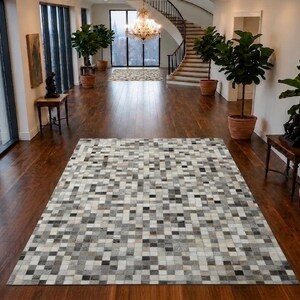 May include: A rectangular area rug featuring a mosaic pattern of small square tiles in varying shades of gray, white, and black. The rug is laid flat on a dark wooden floor, displaying a consistent grid-like arrangement of neutral-toned pieces.