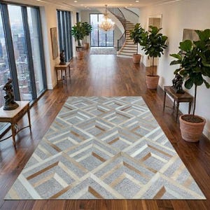 Op de afbeelding: Een rechthoekig vloerkleed met een geometrisch patroon van in elkaar grijpende lichtgrijze, witte en beige ruitvormen. Het kleed ligt op een donkerbruine houten vloer in een ruime hal met grote ramen, kamerplanten en een trap.