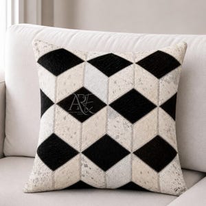 May include: A square throw pillow featuring a geometric pattern of black and speckled off-white diamond shapes. The surface is composed of stitched panels with a textured hair-on-hide appearance. A black logo with the letters AE is visible.