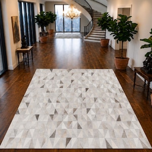 Puede incluir: Una alfombra rectangular con un patrón geométrico de triángulos en varios tonos de blanco, gris claro y gris oscuro. La alfombra está compuesta por numerosos paneles triangulares pequeños cosidos, creando una superficie texturizada.