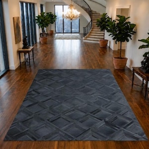 May include: A rectangular area rug featuring a dark gray geometric pattern of interlocking squares and rectangles. The rug is positioned centrally on a polished wooden floor within a spacious room that includes potted plants and furniture.