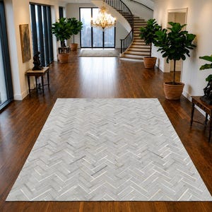 Puede incluir: Una alfombra rectangular con un patrón de espiga en gris claro y blanco. La alfombra está colocada en el centro sobre un suelo de madera dura de color marrón oscuro en una habitación espaciosa con plantas en macetas, mesas y una escalera.